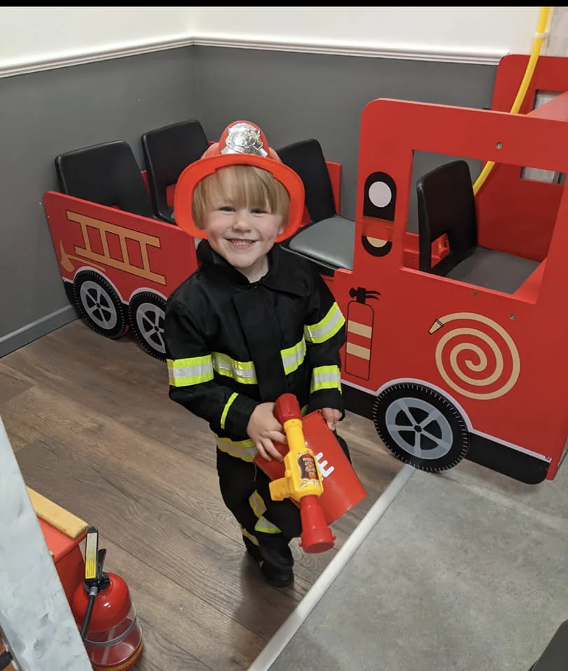 A smiling little firefighter playing beside the red fire engine at Let’s Pretend